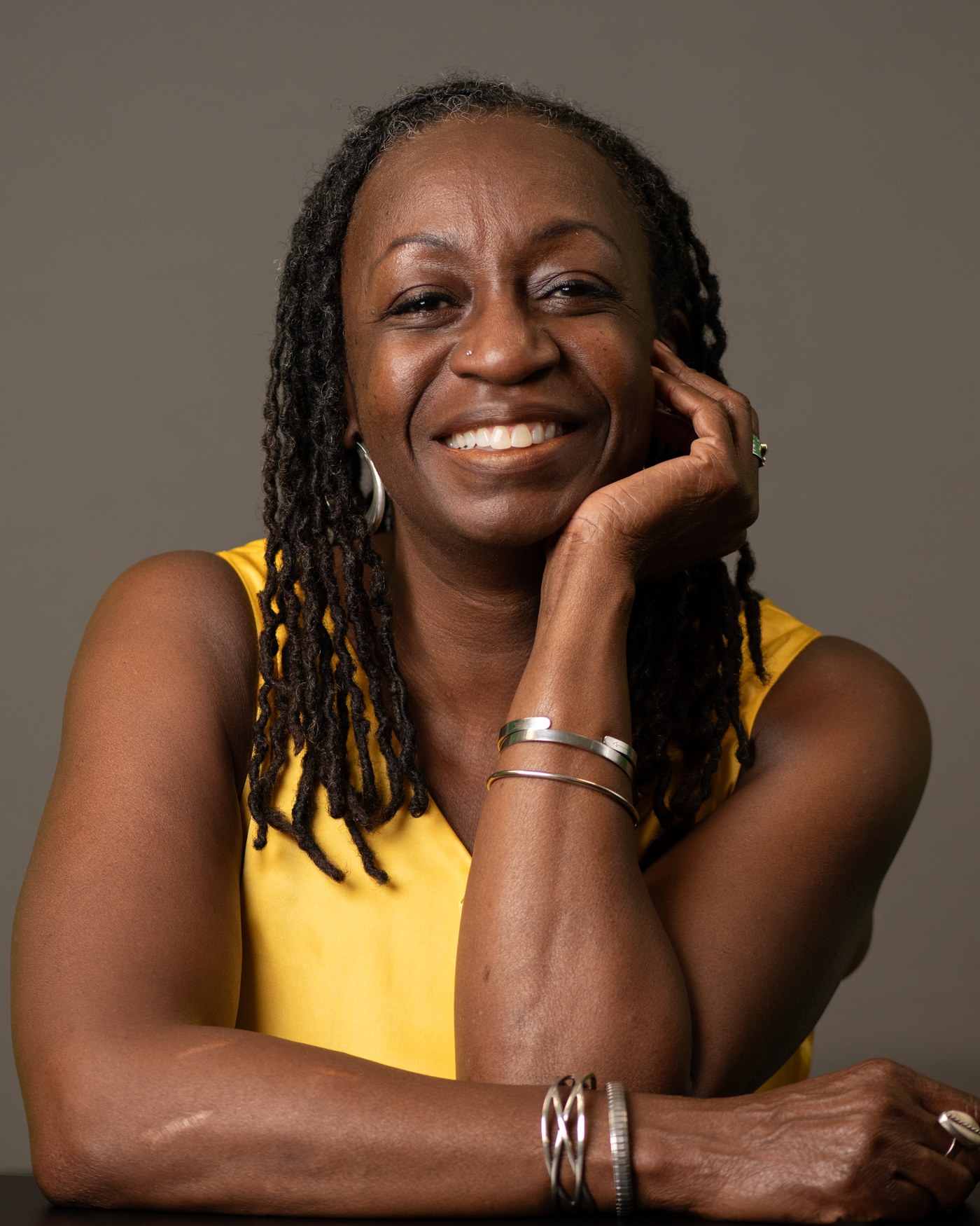 Portrait of Wendy Shaia smiling, with long locs, chin resting on her hand, wearing a yellow top against a gray background
