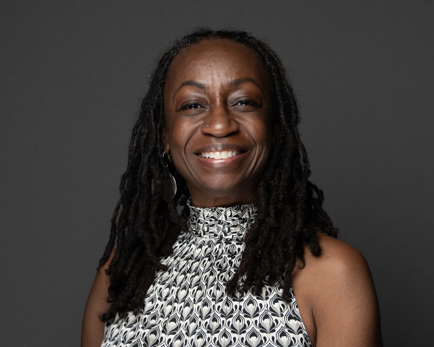 Portrait of Wendy Shaia smiling, with long locs, wearing a black and white patterned top against a dark gray background